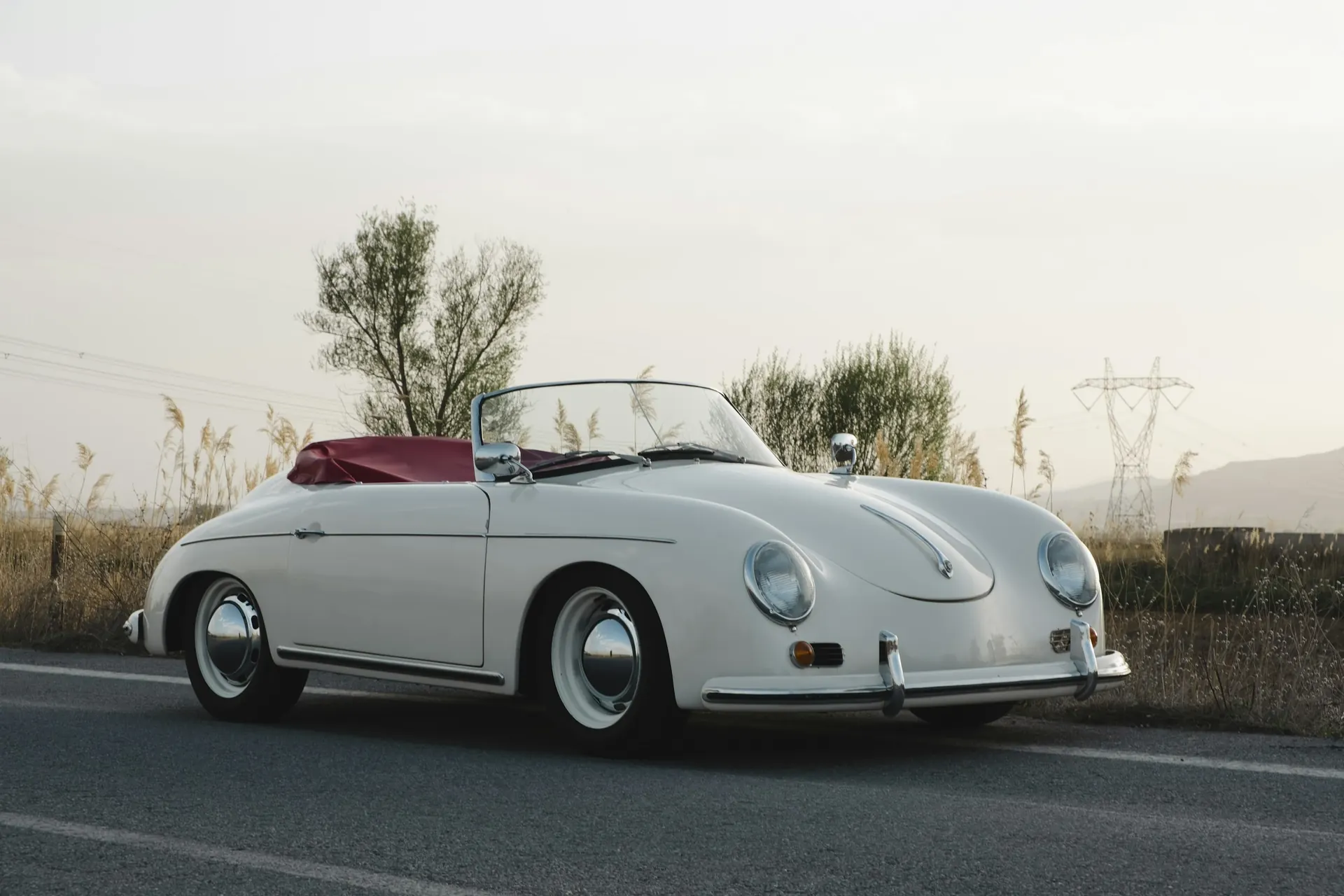 A white Porsche 356 Speedster with a red tonneau cover, parked on a country road in evening light.