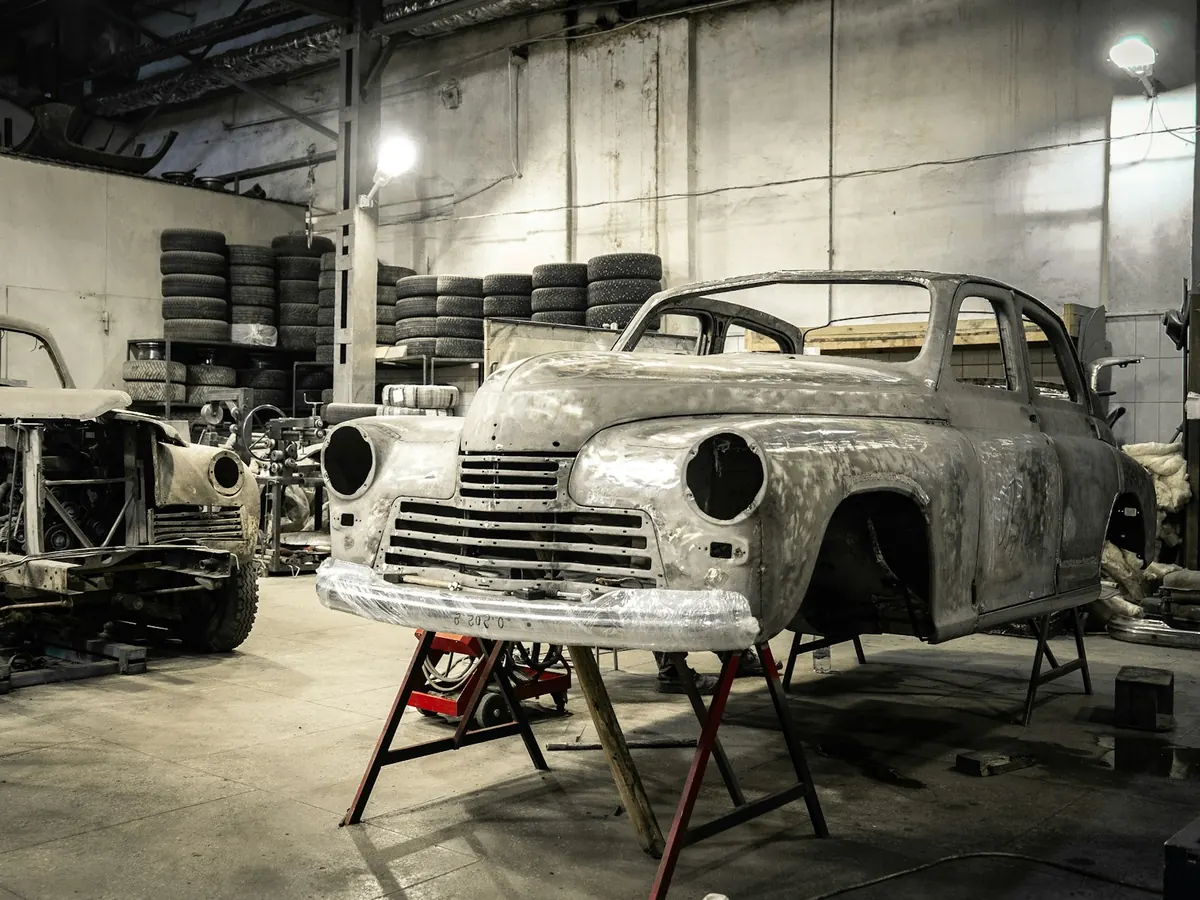 Two classic car bodies stripped to bare metal in a workshop, one on red stands, with stacked tyres behind.